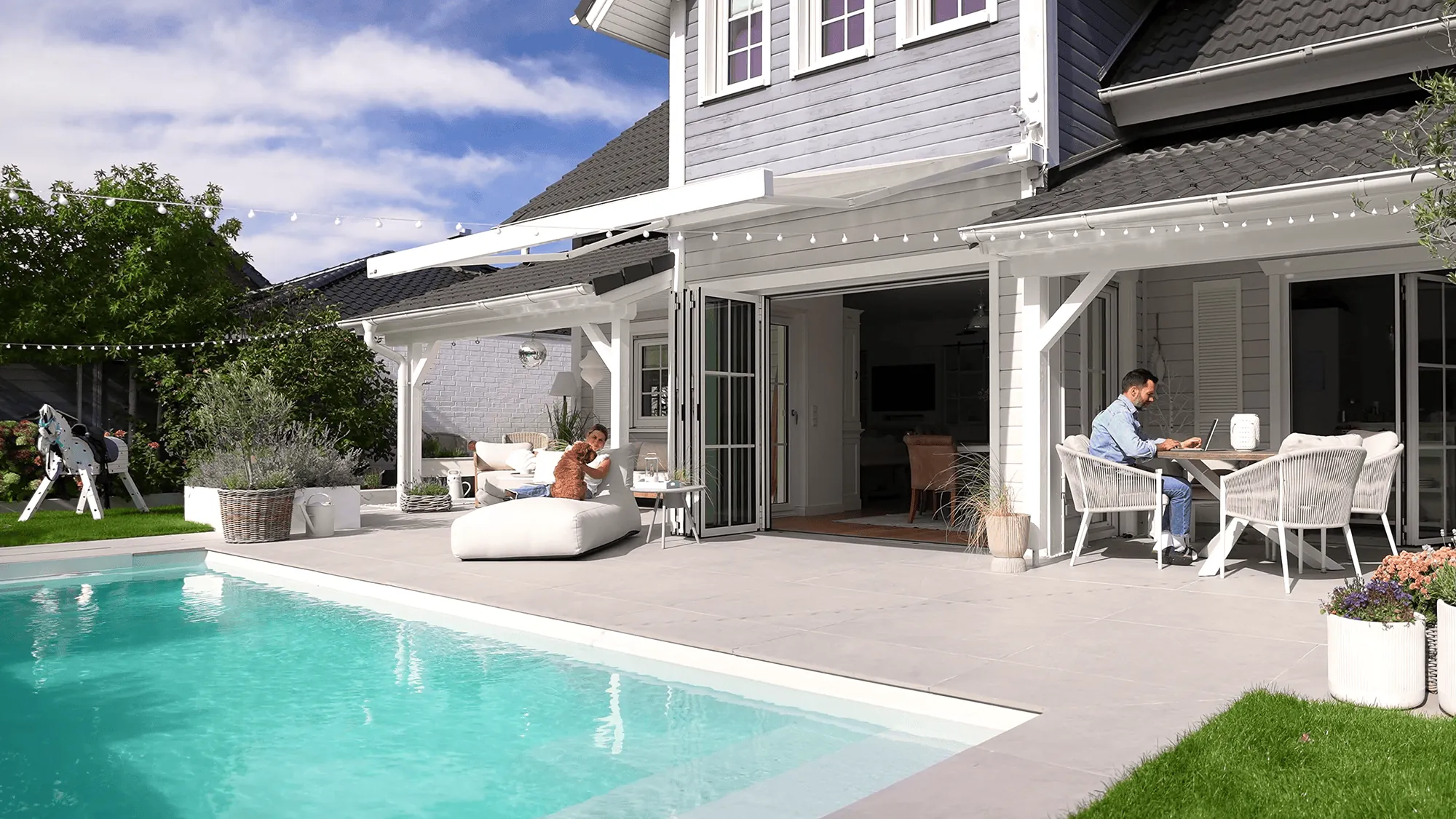 Da ihr Pool direkt an die Terrasse angrenzt, wollten die beiden Eigenheimbesitzer den Bereich offen und großzügig gestalten und entschieden sich bewusst gegen eine fest installierte Pergola. Die Wahl fiel daher auf die Gelenkarmmarkise markilux 970 mit Vollkassette, die ohne Stützpfosten auskommt und an der Hauswand montiert wird. Bei Nichtgebrauch lässt sie sich vollständig einfahren, sodass der Blick in den Garten und auf den Pool frei bleibt. Mit vier Metern Breite und drei Metern Ausfall deckt sie den Terrassenbereich großzügig ab. Das Gestell in Verkehrsweiß (RAL 9016) greift die weißen Elemente der Fassade auf und harmonisiert mit der grauen Holzfassade. Das Tuch, ebenso ein weißer Farbton, fügt sich zurückhaltend in das Gesamtbild ein.